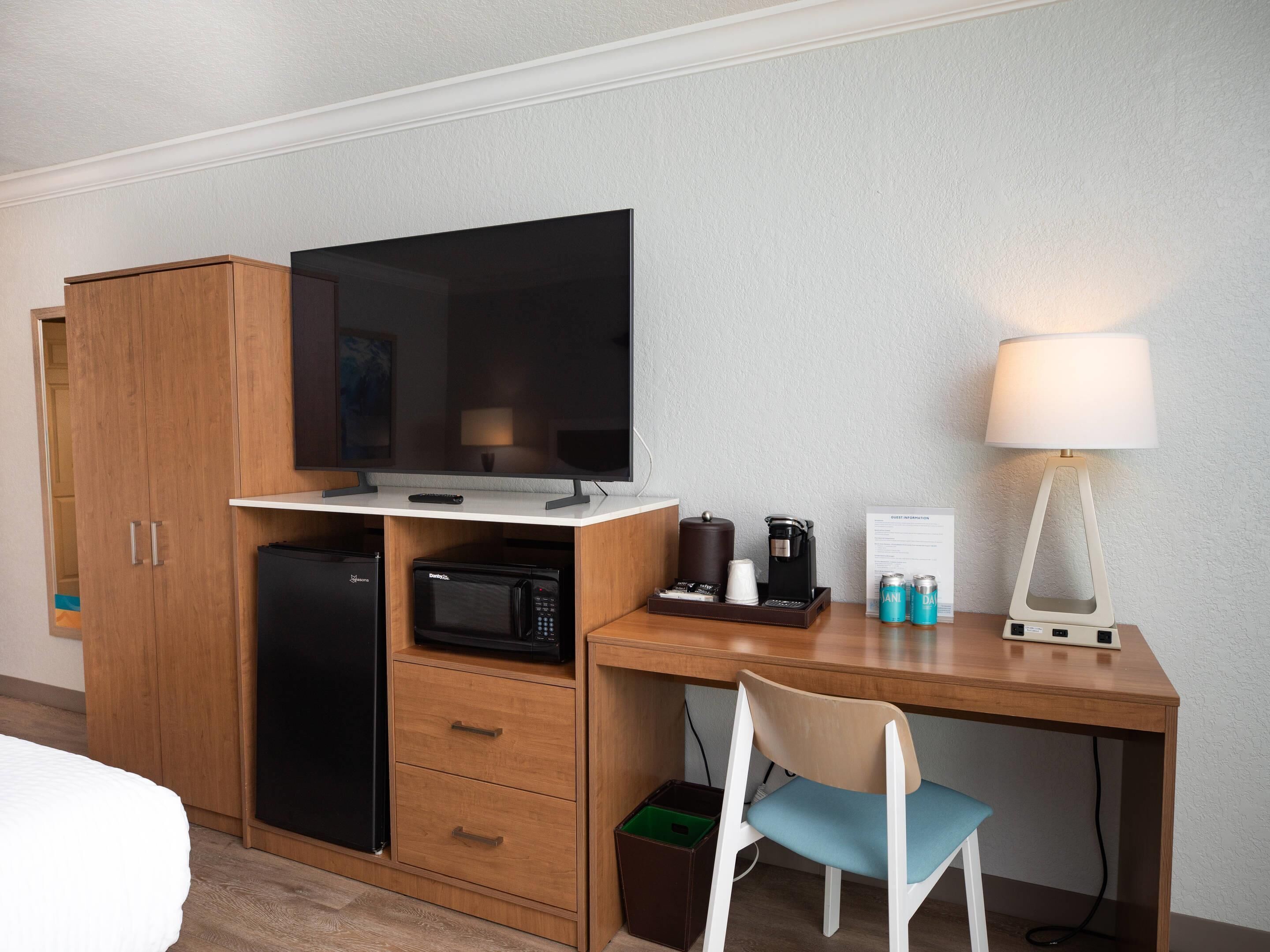 Habitación de hotel con escritorio de madera, televisor, microondas, cafetera, lámpara y una silla.