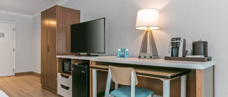 A compact hotel room with a kitchenette, a flat-screen TV, a desk with a lamp, a chair, and a coffee maker.