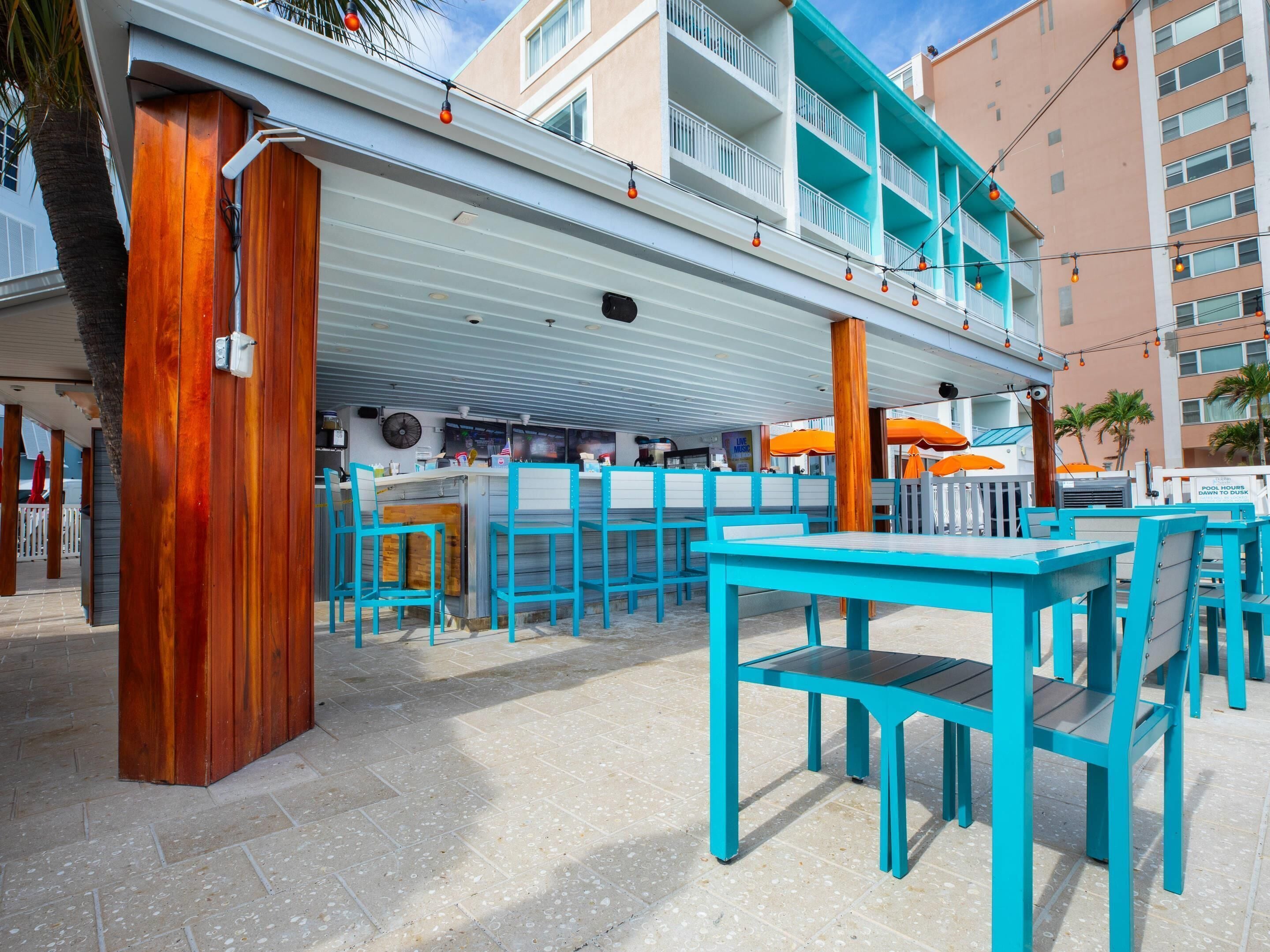 Un bar en la playa con mesas al aire libre, un área de piscina y un edificio con balcones.