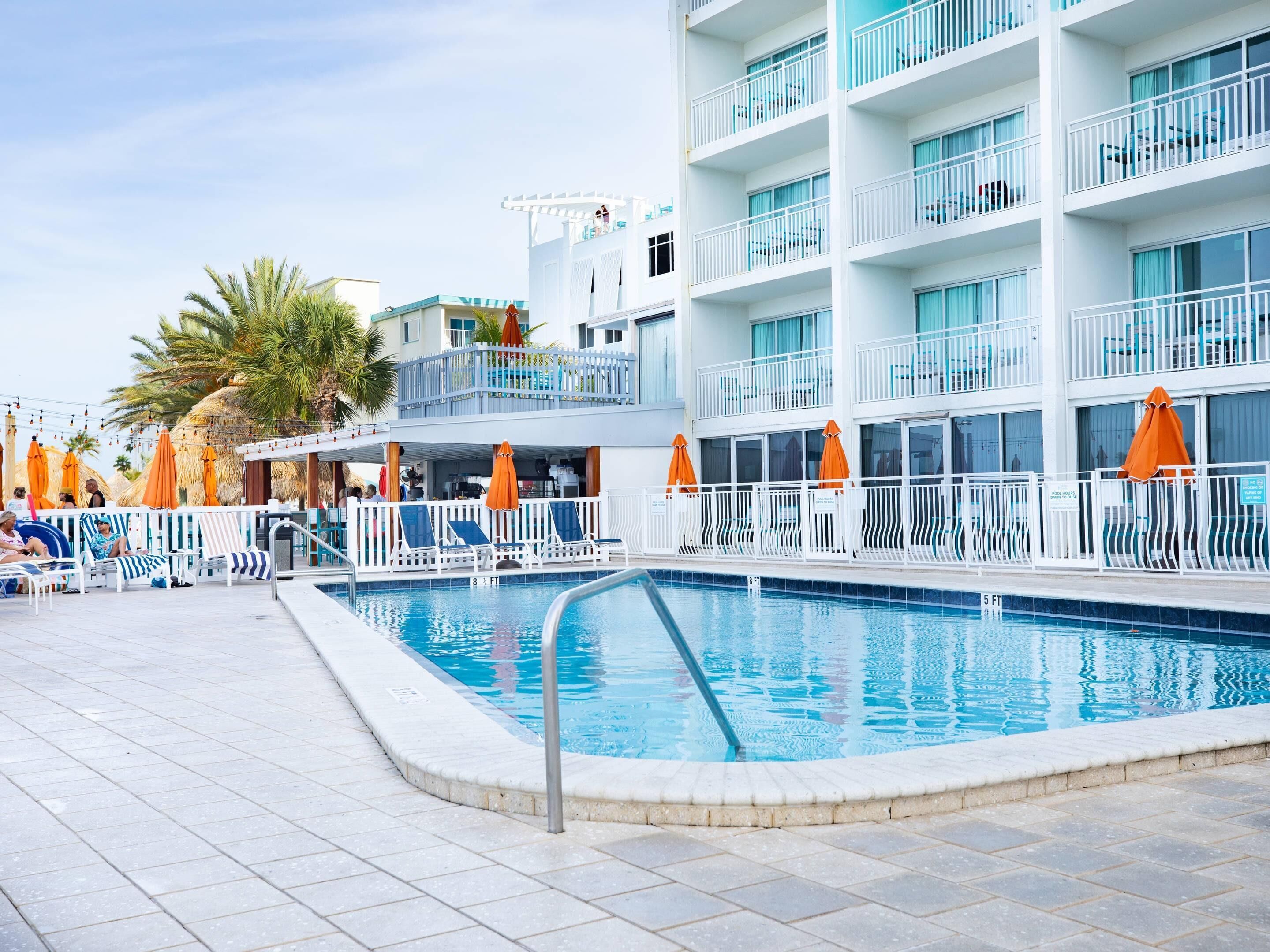 Una piscina con sillones y sombrillas, un edificio de varios pisos con balcones y palmeras.
