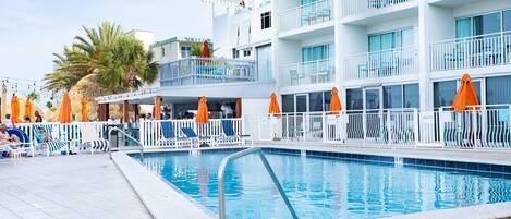 Una piscina con sillones y sombrillas, un edificio de varios pisos con balcones y palmeras.