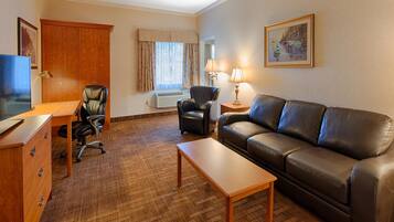 A hotel room with a wooden desk, a leather sofa, a leather armchair, a small table, a TV, a lamp, a window with curtains, and a framed picture on the wall.