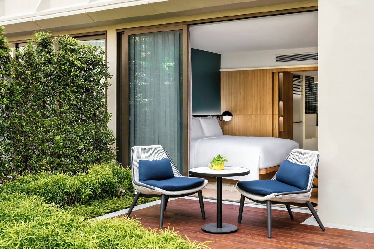 Une chambre d’hôtel moderne avec un grand lit, deux fauteuils en osier, une petite table ronde et une vue sur la verdure environnante.
