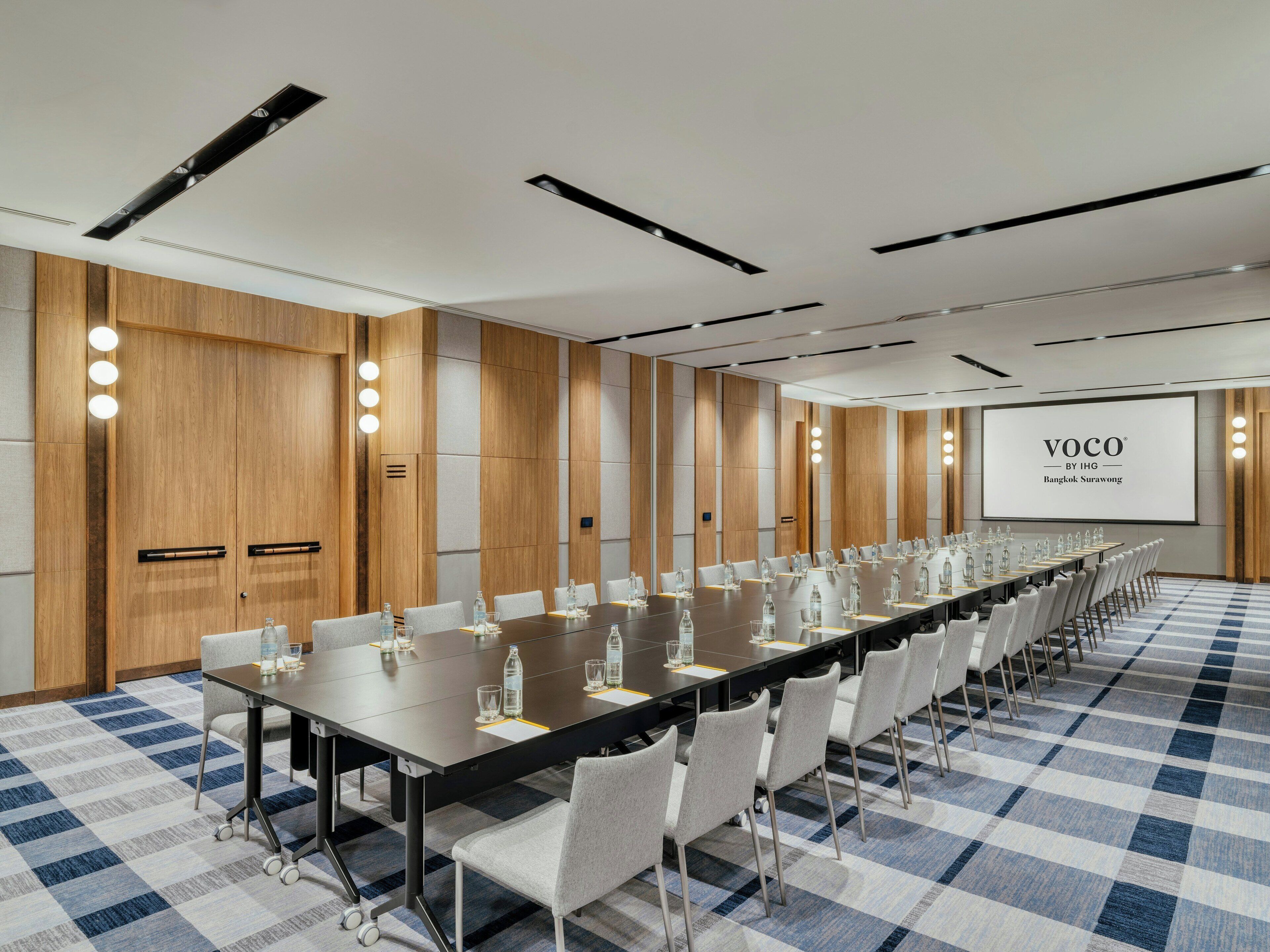 Un salón de conferencias con una larga mesa, sillas y una pantalla mostrando 'VOCO BY IHG Bangkok Surawong'.