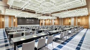 Een vergaderzaal met een groot scherm waarop 'VOCO BY IHG Bangkok Surawong' staat. Er staan rijen tafels met stoelen, waterflessen en naambadges klaar voor een vergadering.