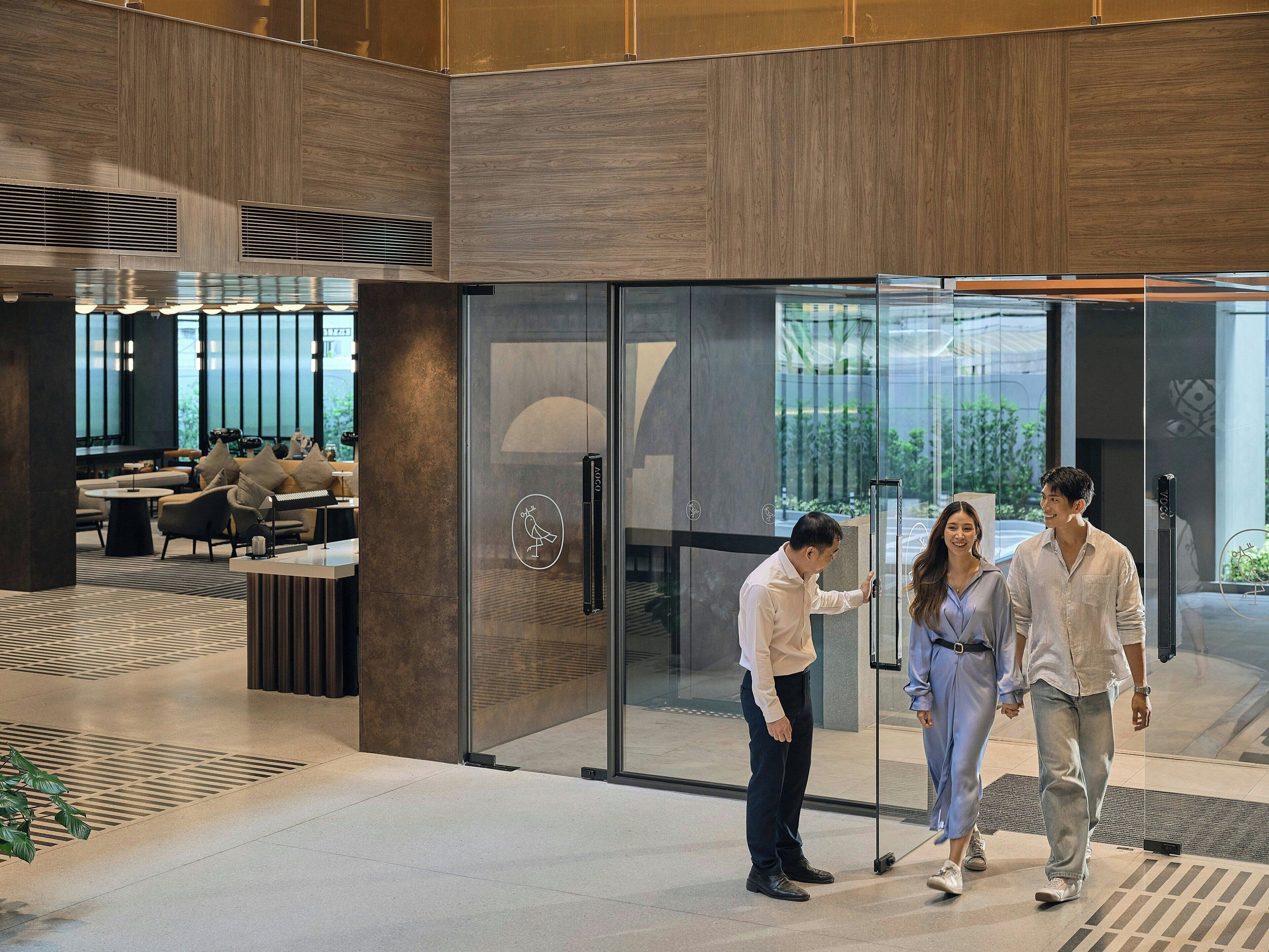A modern interior with a glass door entrance, a seating area with tables and chairs, and a view of greenery outside.