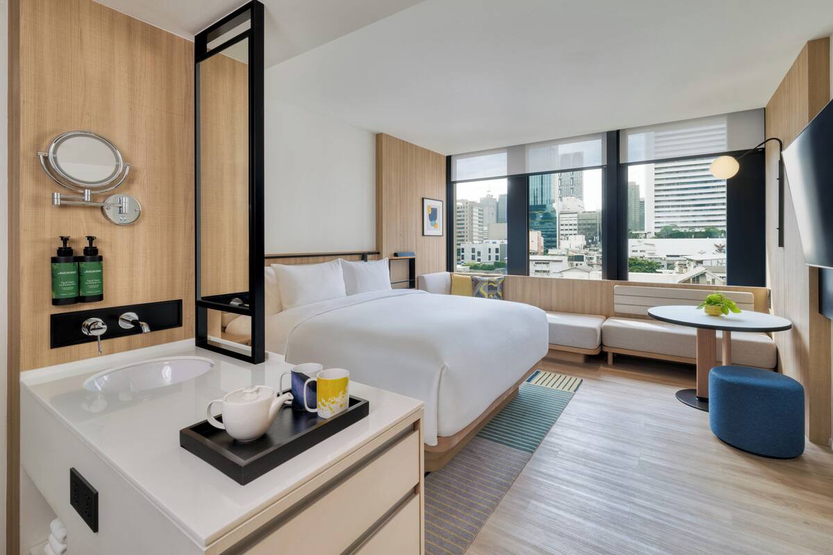 Une chambre d’hôtel moderne dotée d’un grand lit, d’un coin salon avec vue, d’une salle de bain avec miroir et articles de toilette, et offrant une vue sur la ville grâce à de grandes fenêtres.
