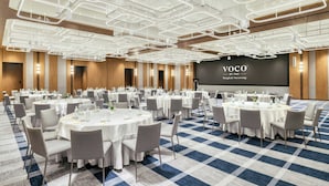 Un salón de conferencias con varias mesas redondas, sillas grises y una pantalla grande que muestra 'VOCO BY IHG Bangkok Surawong'.