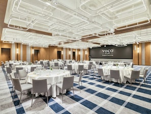 Une salle de conférence dotée de plusieurs tables rondes, de chaises grises et d’un grand écran affichant la mention « VOCO BY IHG Bangkok Surawong ».