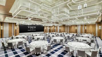 Una sala de conferències amb diverses taules rodones, cadascuna coberta amb mantells blancs i amb caires. Es projecta una imatge gran amb la inscripció 'VOCO BY IHG Bangkok Surawong'.