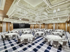 Un salón de conferencias con varias mesas redondas, cada una con mantel blanco y sillas, y una gran pantalla mostrando 'VOCO BY IHG Bangkok Surawong'.