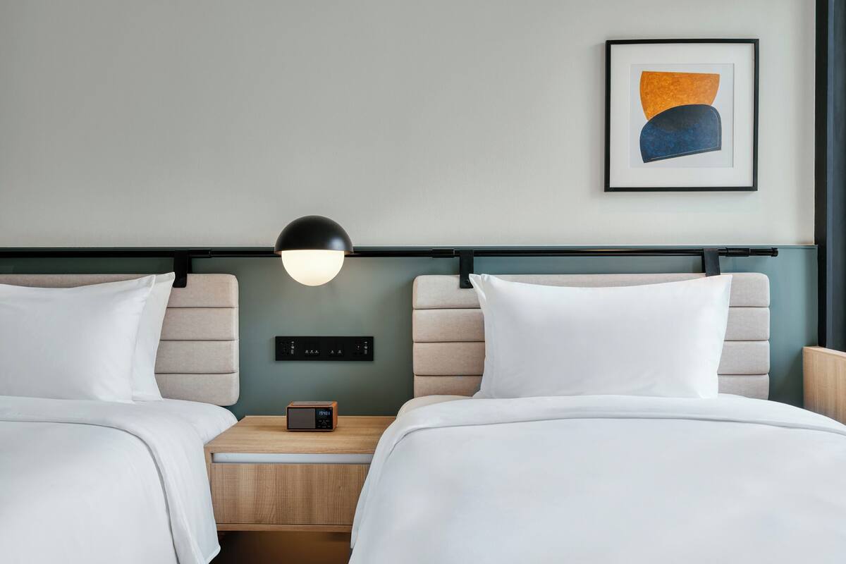 Une chambre d’hôtel avec deux lits, une table de chevet avec une radio, une lampe fixée au mur et un tableau abstrait encadré.