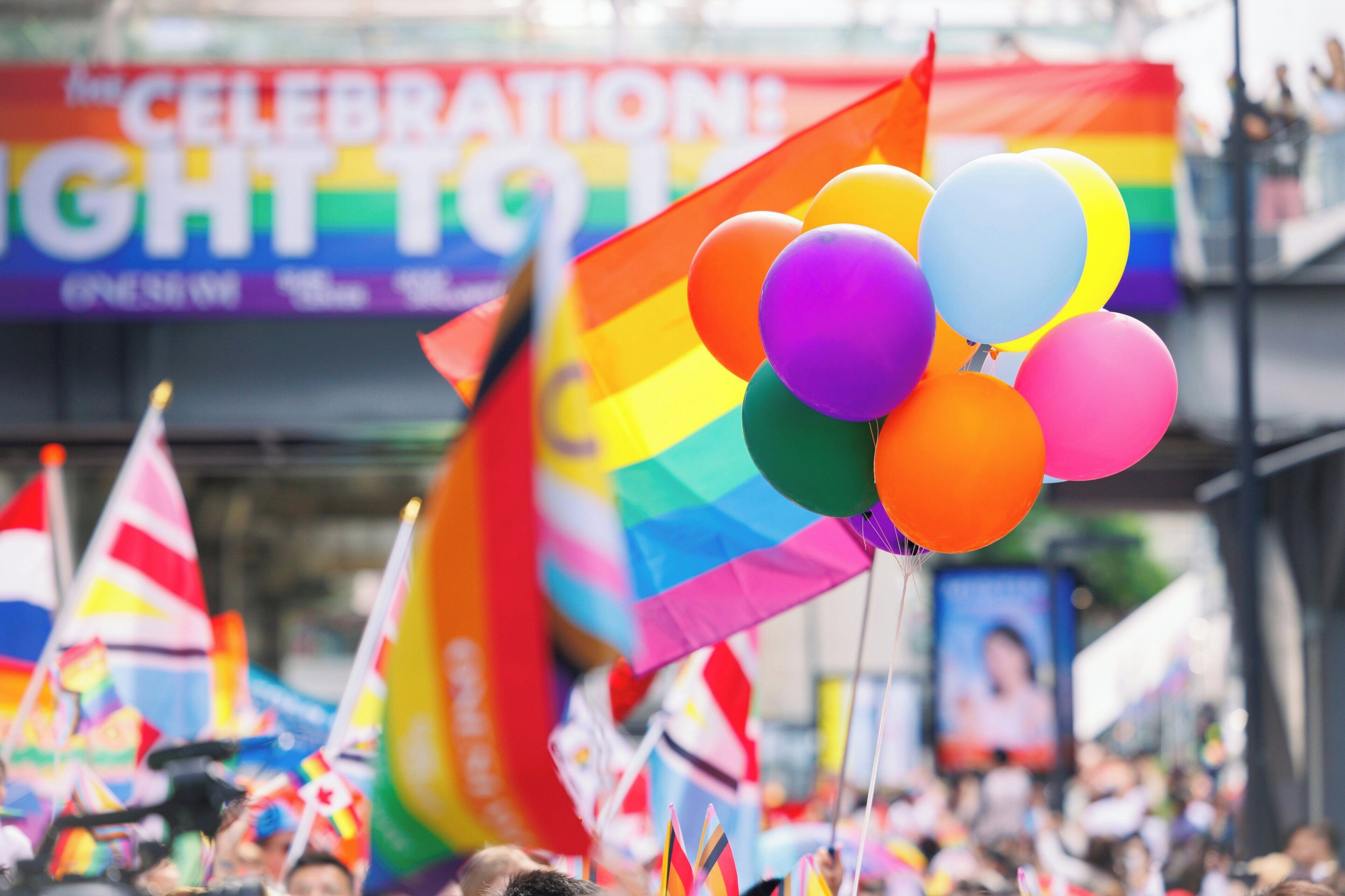 A crowd at a public event with a rainbow flag and balloons.