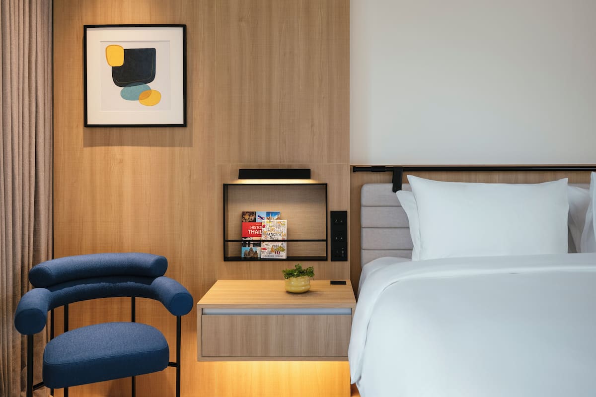Una habitación de hotel moderna con cama, mesita de noche, silla, estante con libros y una obra de arte abstracta enmarcada.
