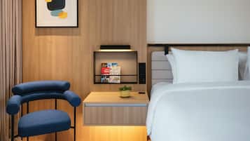 Une chambre d’hôtel moderne, équipée d’un lit, d’une table de chevet, d’une chaise, d’une étagère avec des livres et d’une œuvre d’art abstraite encadrée.
