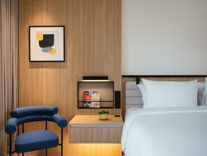 Een moderne hotelkamer met een bed, een nachtkastje, een stoel, een plank met boeken en een lijst met abstracte kunst.