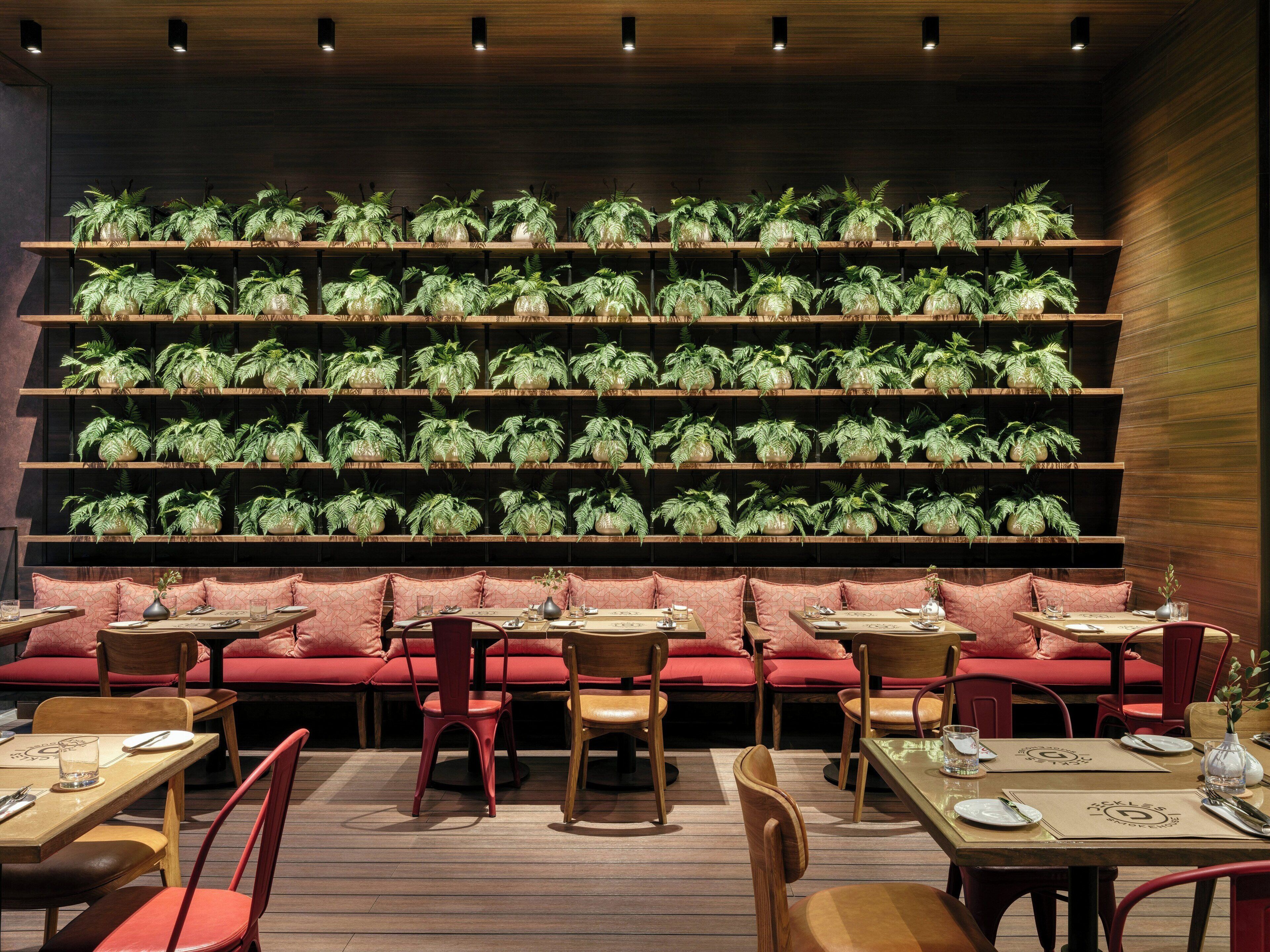 L’intérieur d’un restaurant présente de nombreuses tables et des sièges rembourrés de couleur rouge, le tout étant entouré d’étag ères remplies de plantes vertes.