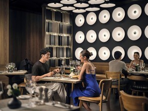 Een restaurantinterieur met ronde tafels, houten stoelen en een muur met ronde lichtarmaturen.