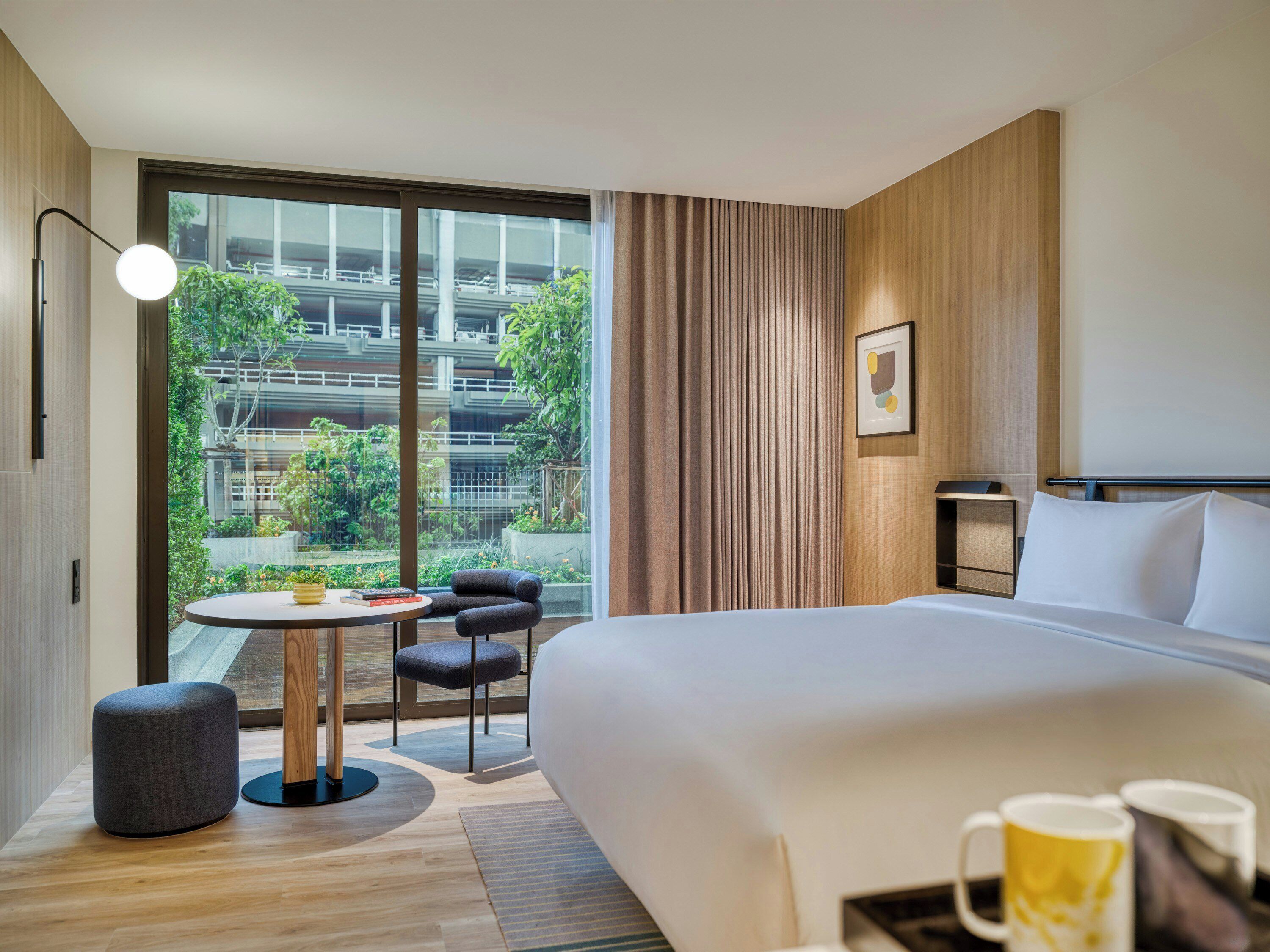 A modern hotel room with a large bed, a round table with chairs, a small stool, and a view of greenery and buildings outside.