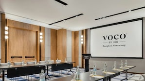 Un salón de conferencias con una larga mesa, sillas y una pantalla que muestra 'VOCO BY IHG Bangkok Surawong'.