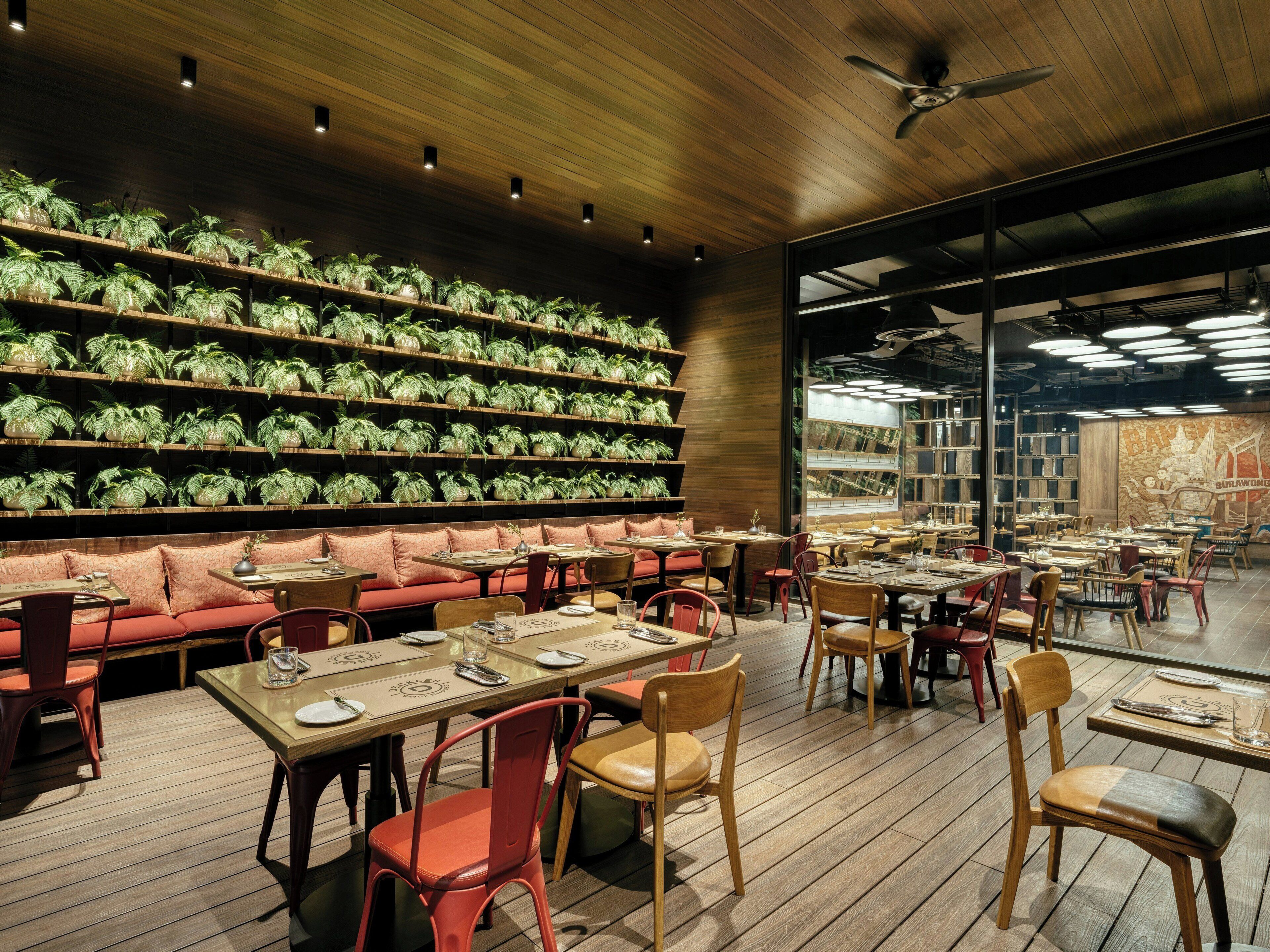 Interior de restaurante con pisos de madera, asientos acolchados rojos y estanterías llenas de plantas verdes.
