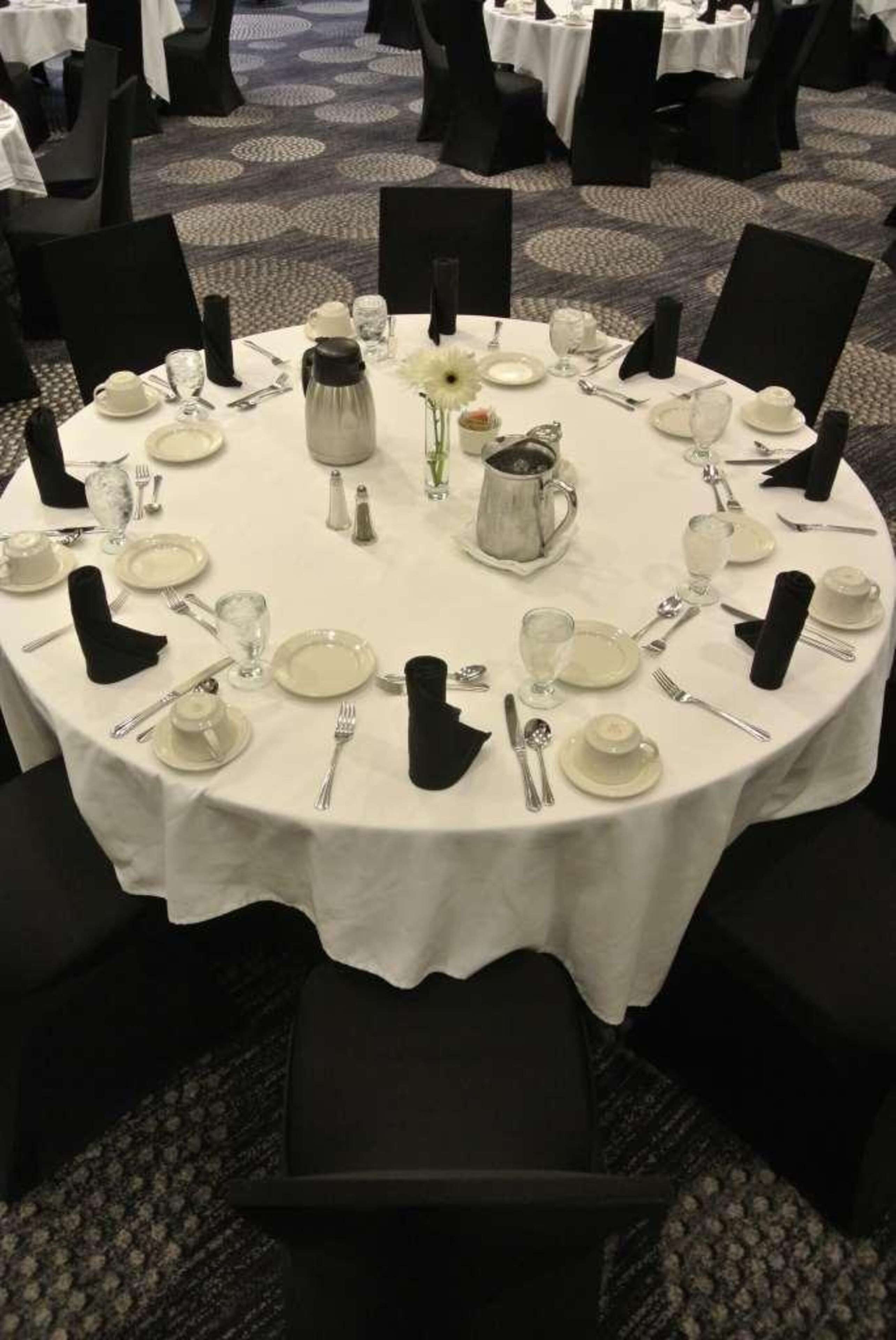Une table ronde dressée pour un événement formel, avec des nappes blanches, des chaises noires, et des couverts et des verres disposés avec soin.