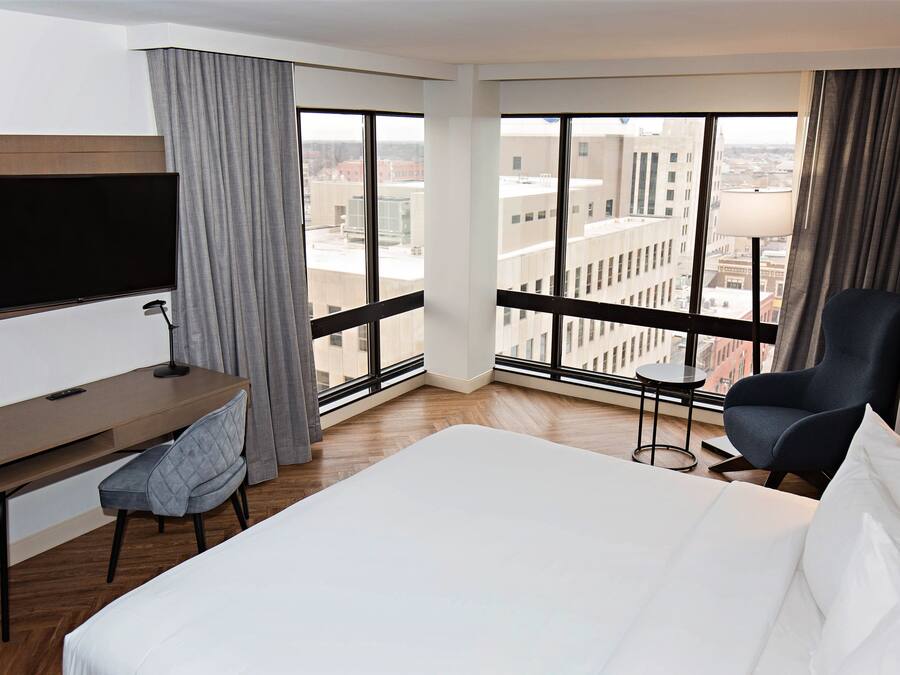 A hotel room with a bed, a desk, a chair, a TV, and large windows offering a city view.