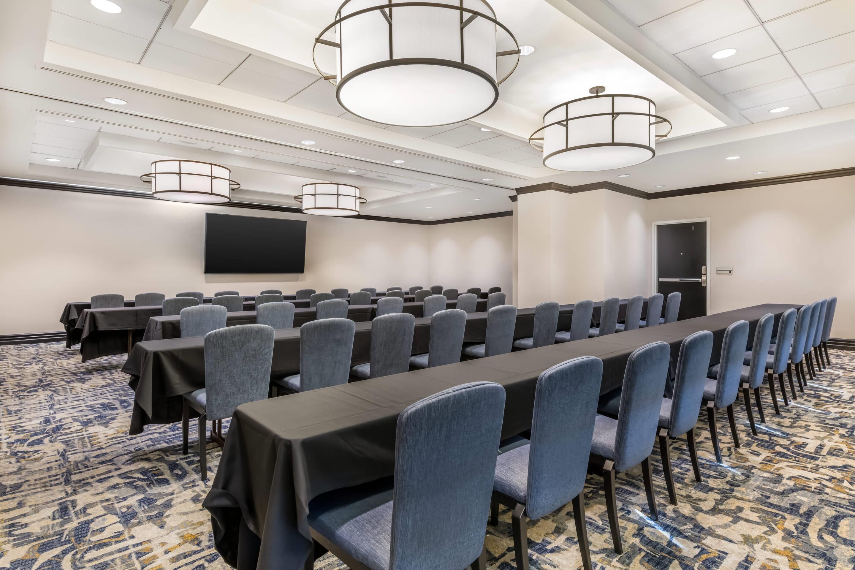 Un salón de conferencias con filas de mesas y sillas, una pantalla grande y una alfombra estampada.