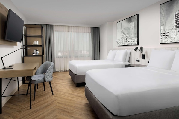 Habitación de hotel con dos camas, un escritorio con una silla, un televisor montado en la pared y obras de arte en las paredes.