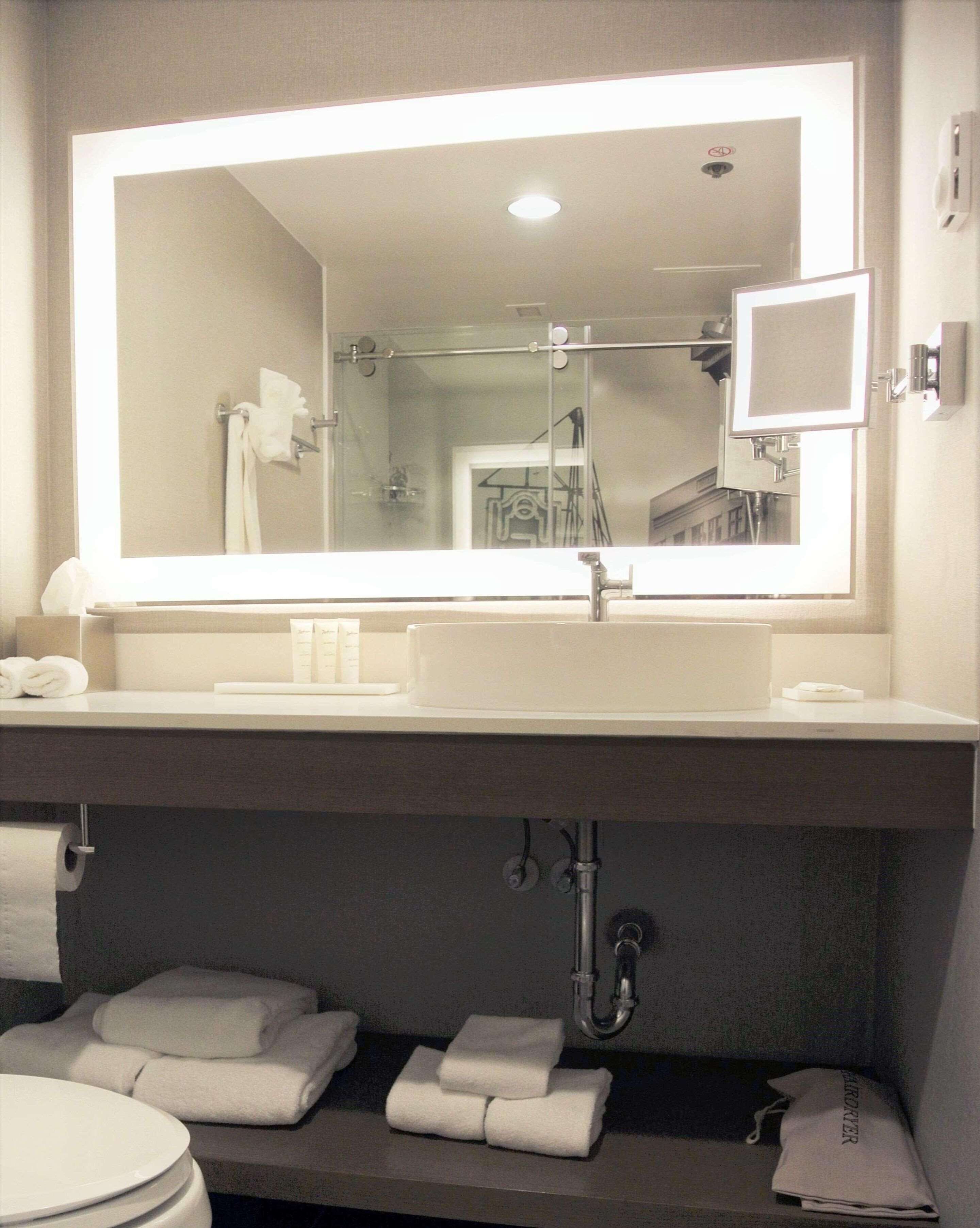A bathroom with a large mirror, a sink, and a toilet. There are towels on the counter and a showerhead above the sink.