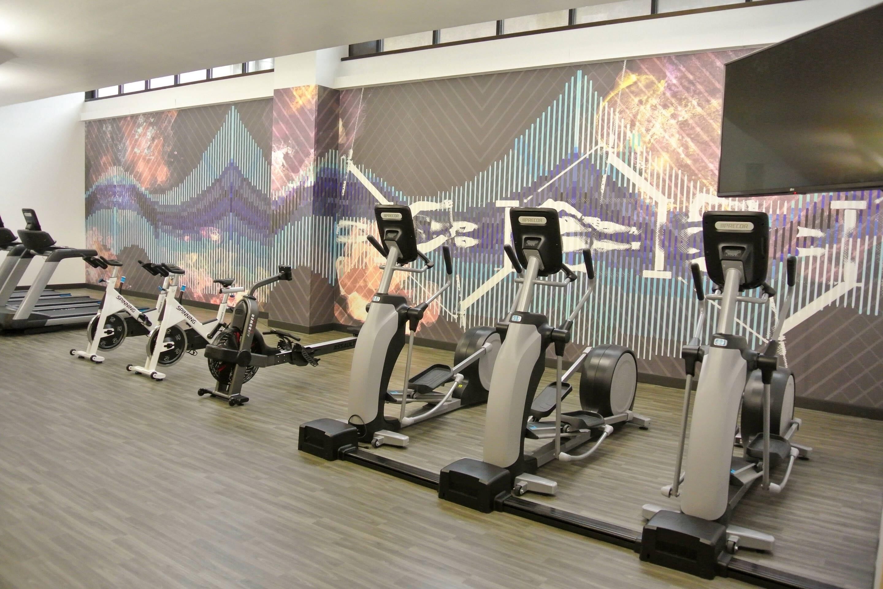 A gym with stationary bikes and ellipticals, a large digital screen, and a wooden floor.