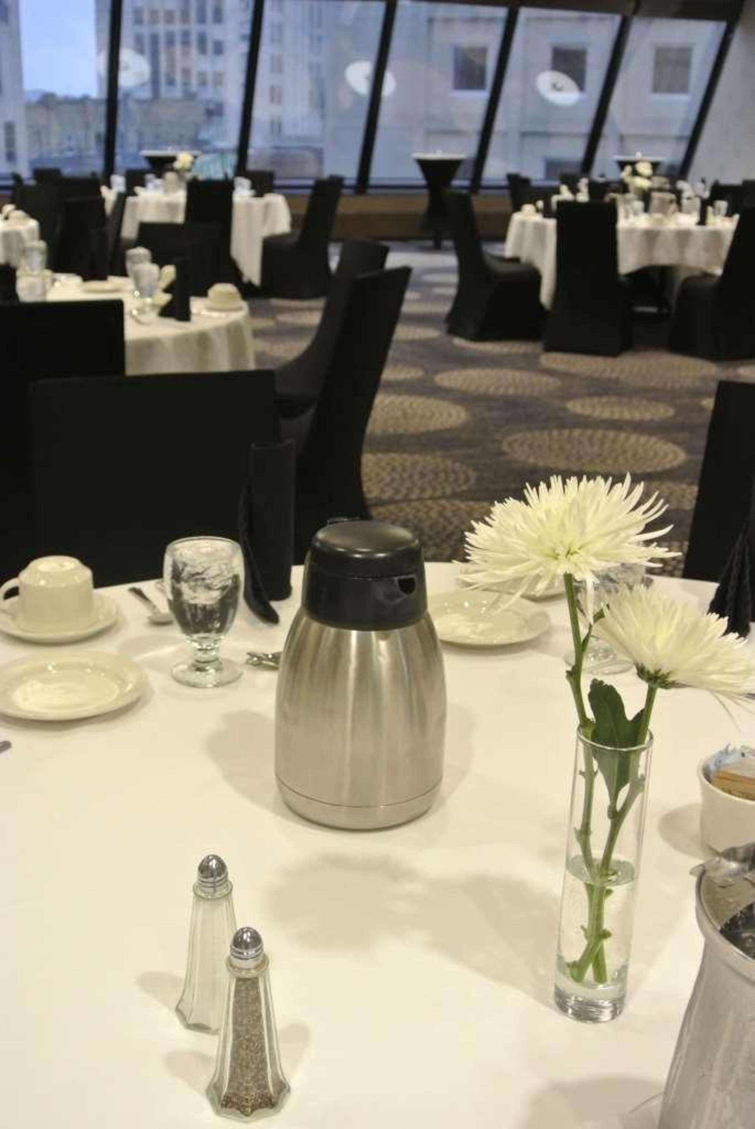 Uma área de refeições com mesas postas, uma cafeteira, saleiro e pimenteiro, e um vaso com flores.