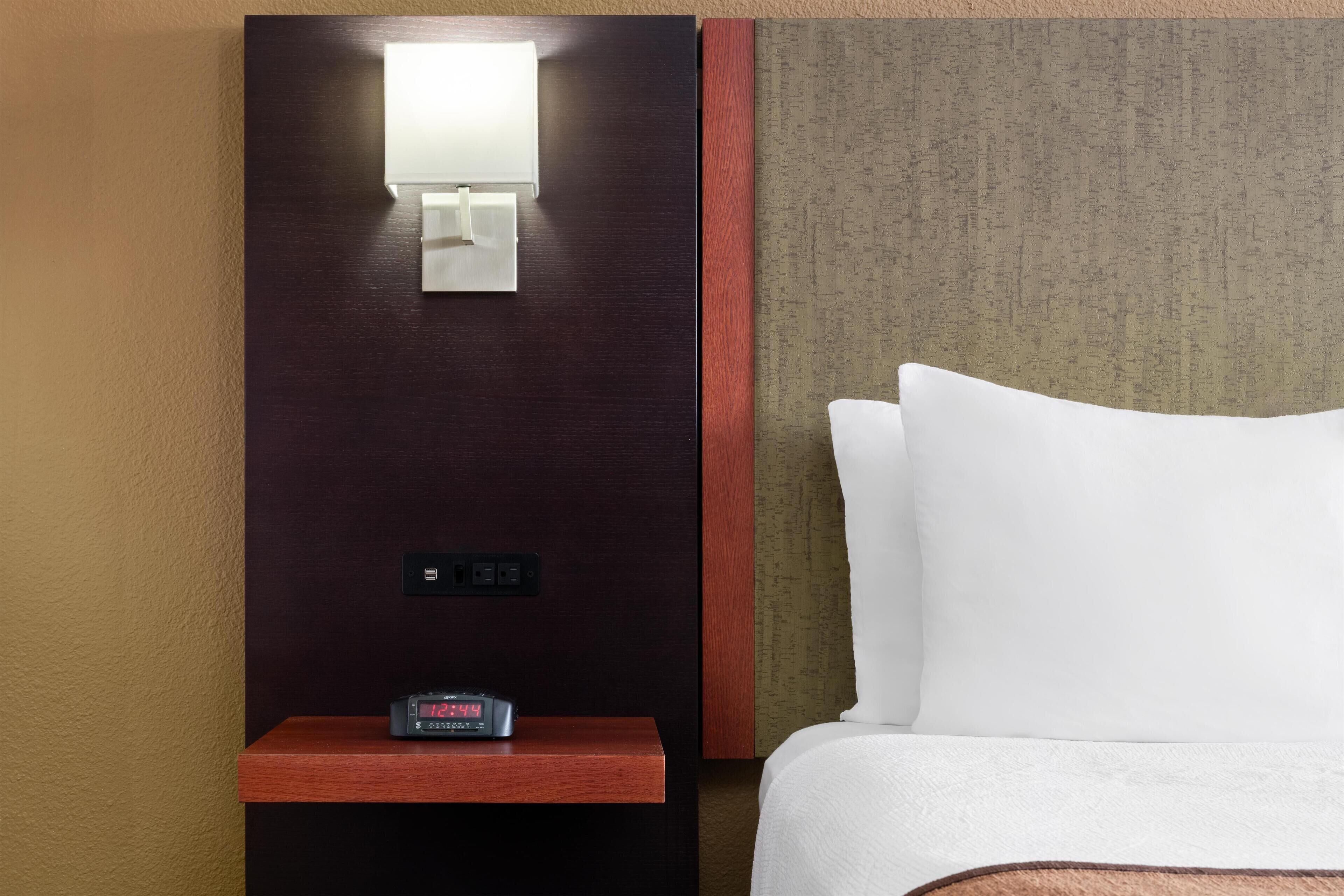 A hotel room with a wooden nightstand, a digital clock, a wall-mounted light, and a bed with white bedding.