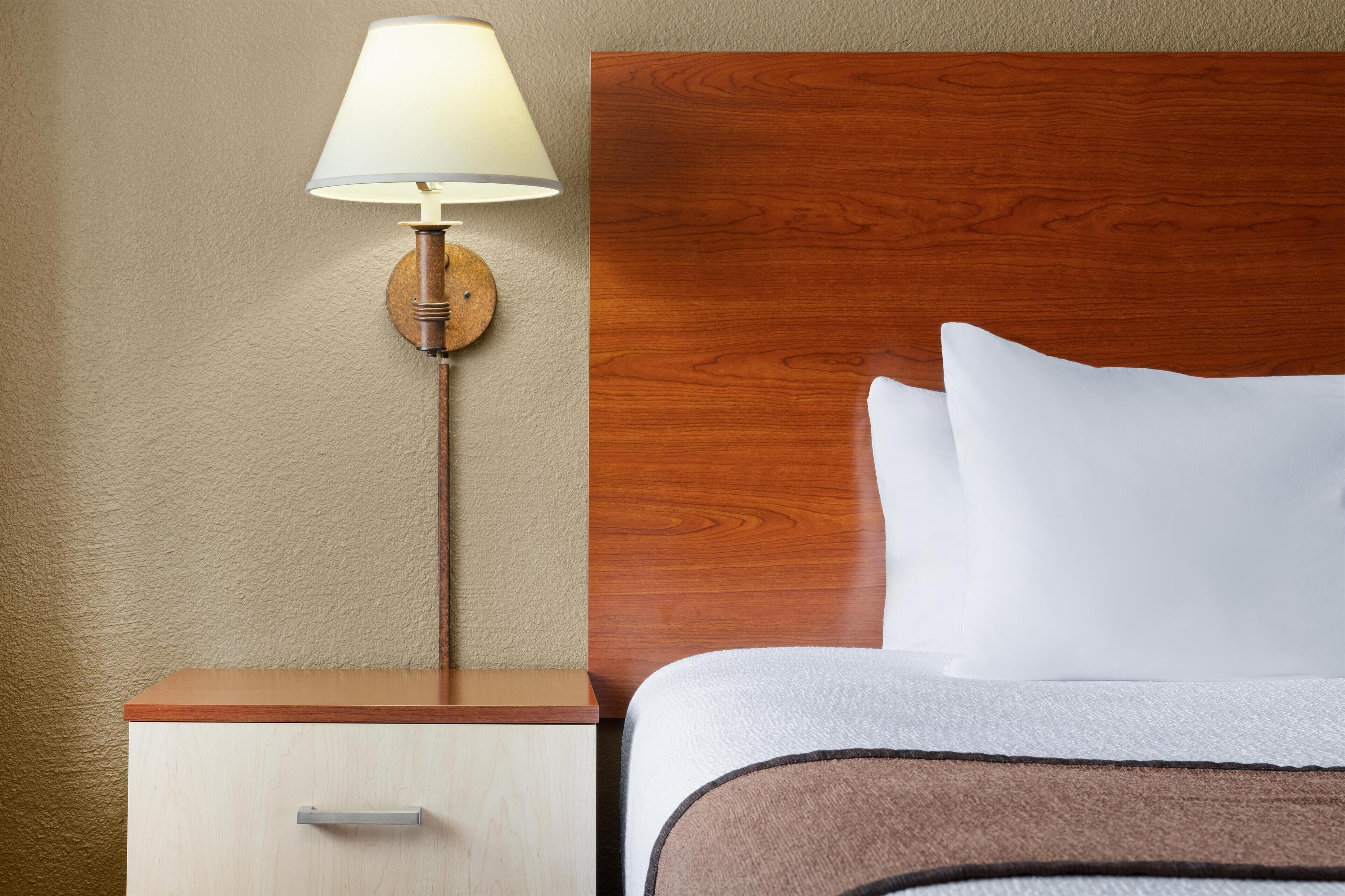 Una cama bien hecha con almohadas blancas y un cabecero de madera, una mesita de noche con una lámpara y un cajón pequeño.