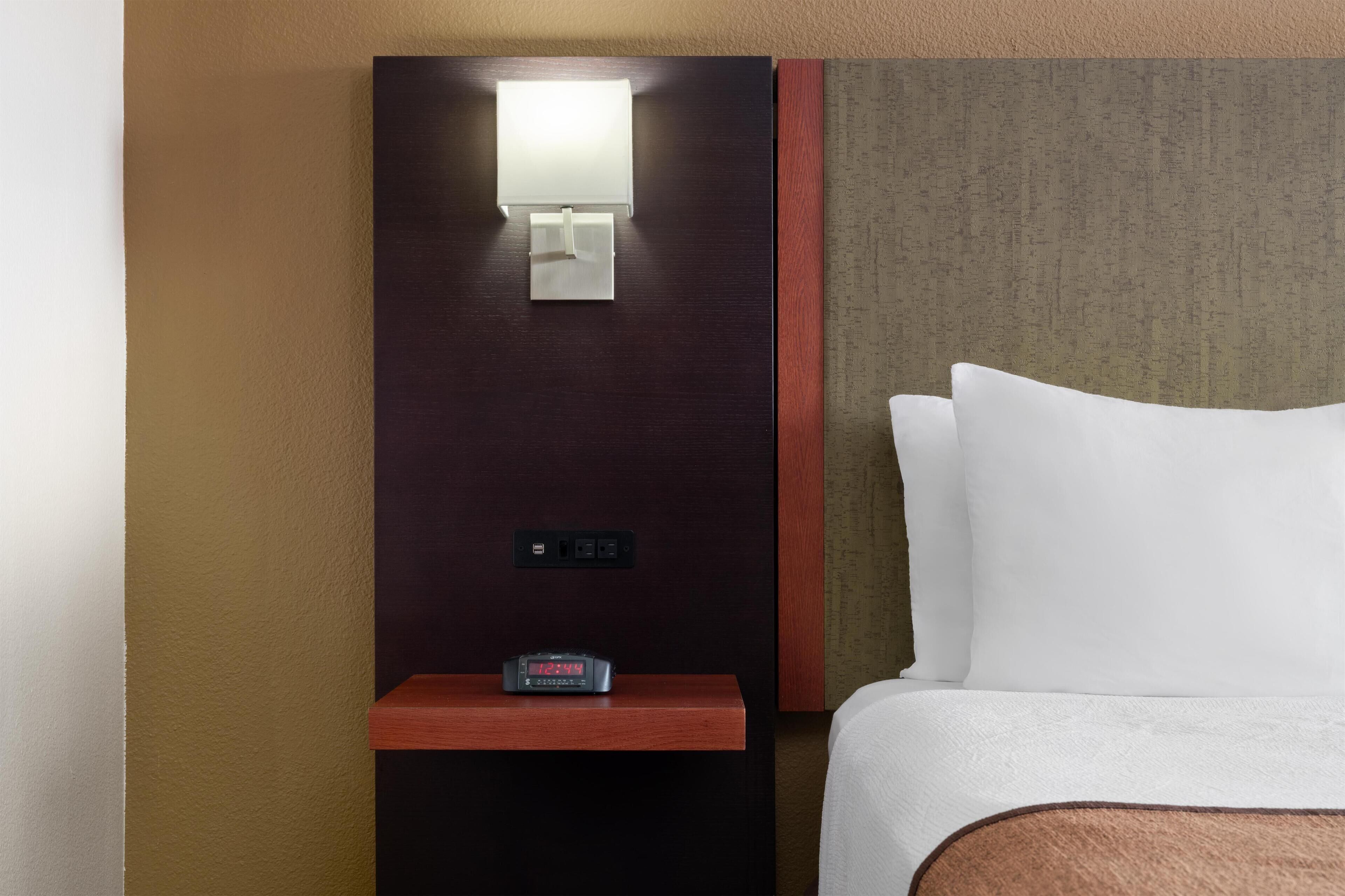 A hotel room with a bed, a nightstand with a clock, a wall-mounted light, and a small shelf.