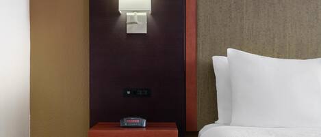 A hotel room with a bed, a nightstand with a clock, a wall-mounted light, and a small shelf.