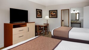 A hotel room with a TV on a wooden cabinet, a desk with a chair, two beds with brown and white bedding, and a bathroom with a mirror and sink.