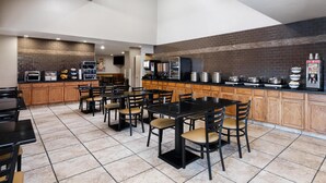 A modern kitchen with a brick backsplash, wooden cabinets, and a central island. There are multiple tables and chairs arranged for dining.