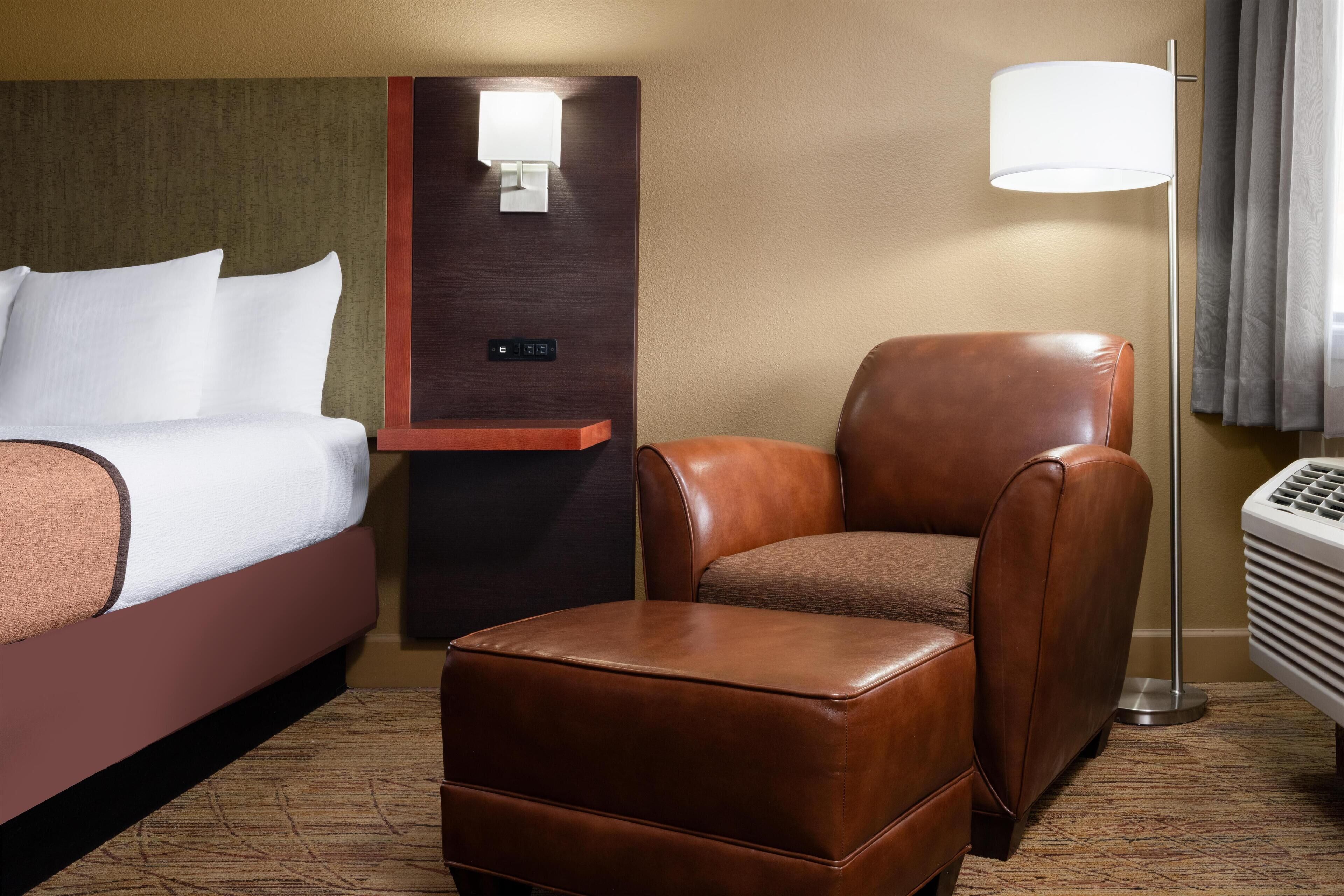 Une chambre d’hôtel avec un lit, un fauteuil en cuir marron, un petit pouf, une lampe fixée au mur et un radiateur portatif.