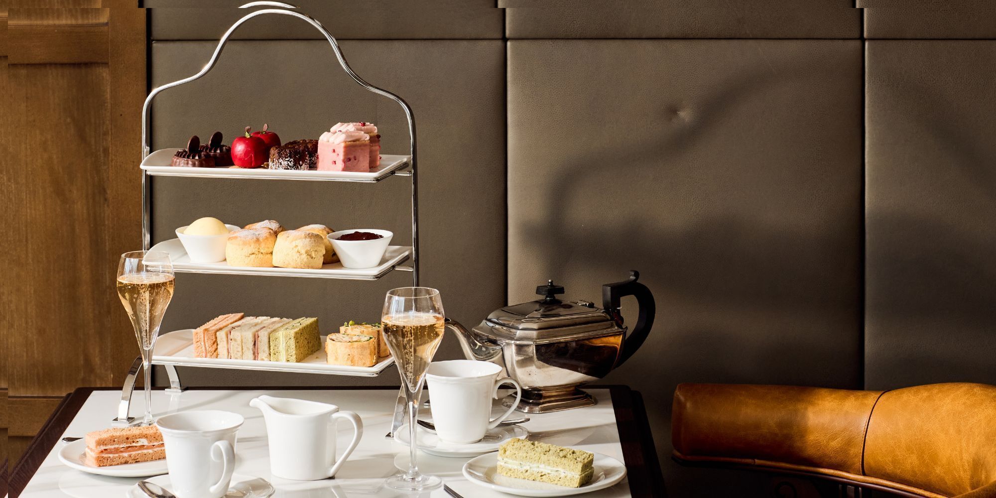 A tiered serving tray with various pastries and a teapot on a table, accompanied by glasses of champagne and a leather chair.