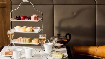 A tiered serving tray with various pastries and a teapot on a table, accompanied by glasses of champagne and a leather chair.