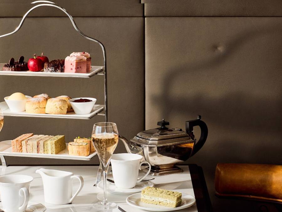 A tiered serving tray with various pastries and a teapot on a table, accompanied by glasses of champagne and a leather chair.
