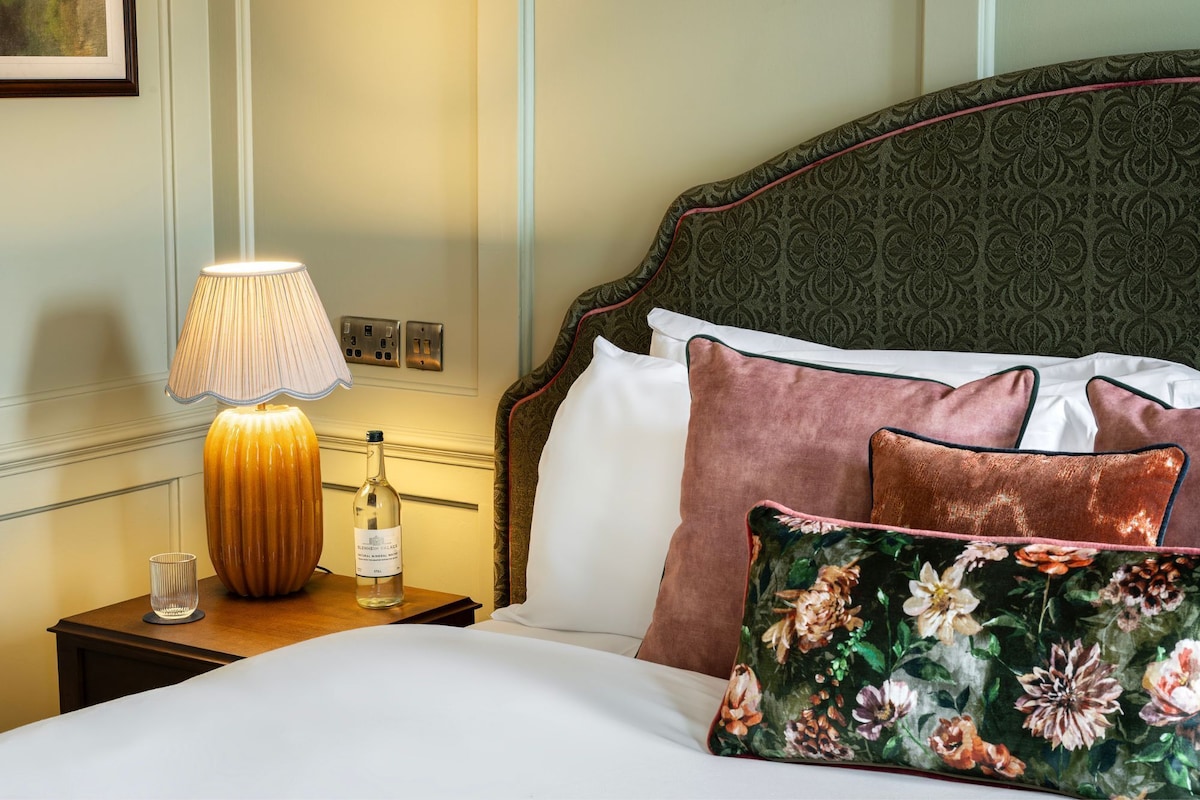 A neatly made bed with floral pillows, a lamp, and a bottle on a bedside table.