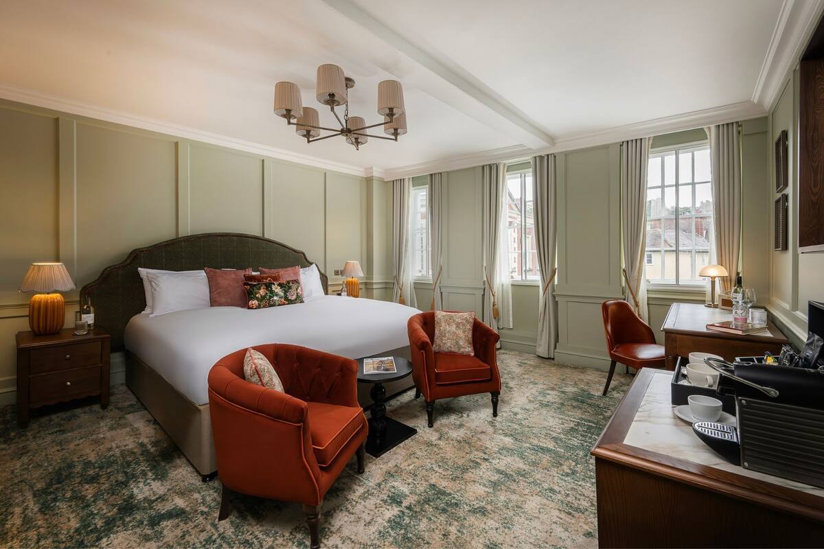 A hotel room with a large bed, two armchairs, a desk with a coffee maker, and a chandelier.