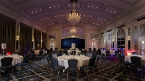 A banquet hall with round tables, chairs, a chandelier, and a stage. - Castle Hotel Windsor (Windsor)