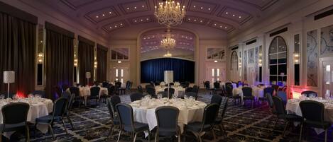 A banquet hall with round tables, chairs, a chandelier, and a stage.