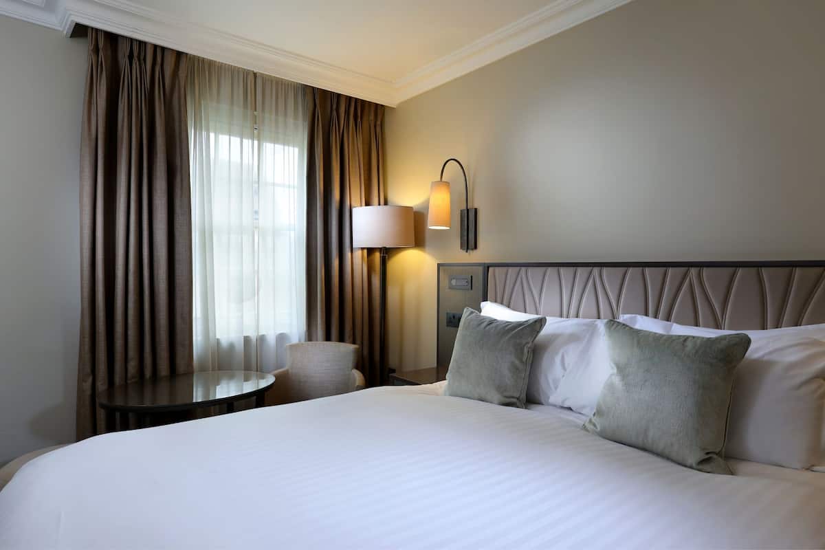A hotel room with a large bed, a round table, a lamp, and a view of the outdoors through sheer curtains.