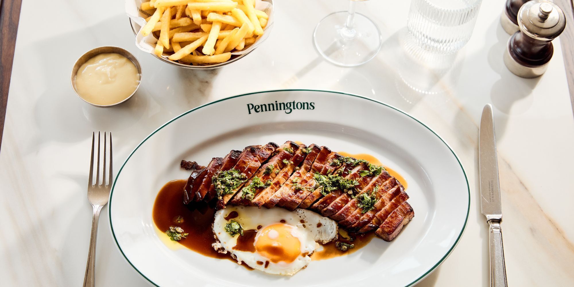A plate of steak with a fried egg and sauce, accompanied by a bowl of fries and a small container of sauce, on a white table with a glass and salt shaker.