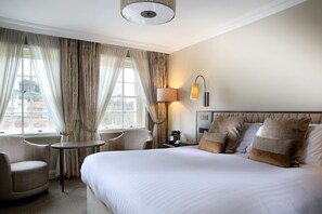 A hotel room with a large bed, two armchairs, a small table, a lamp, and a view of buildings through the window. - Castle Hotel Windsor (Windsor)