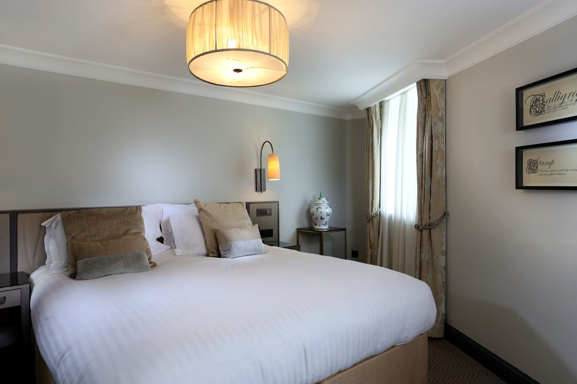 A hotel room with a large bed, a bedside table, a lamp, a vase, and curtains.